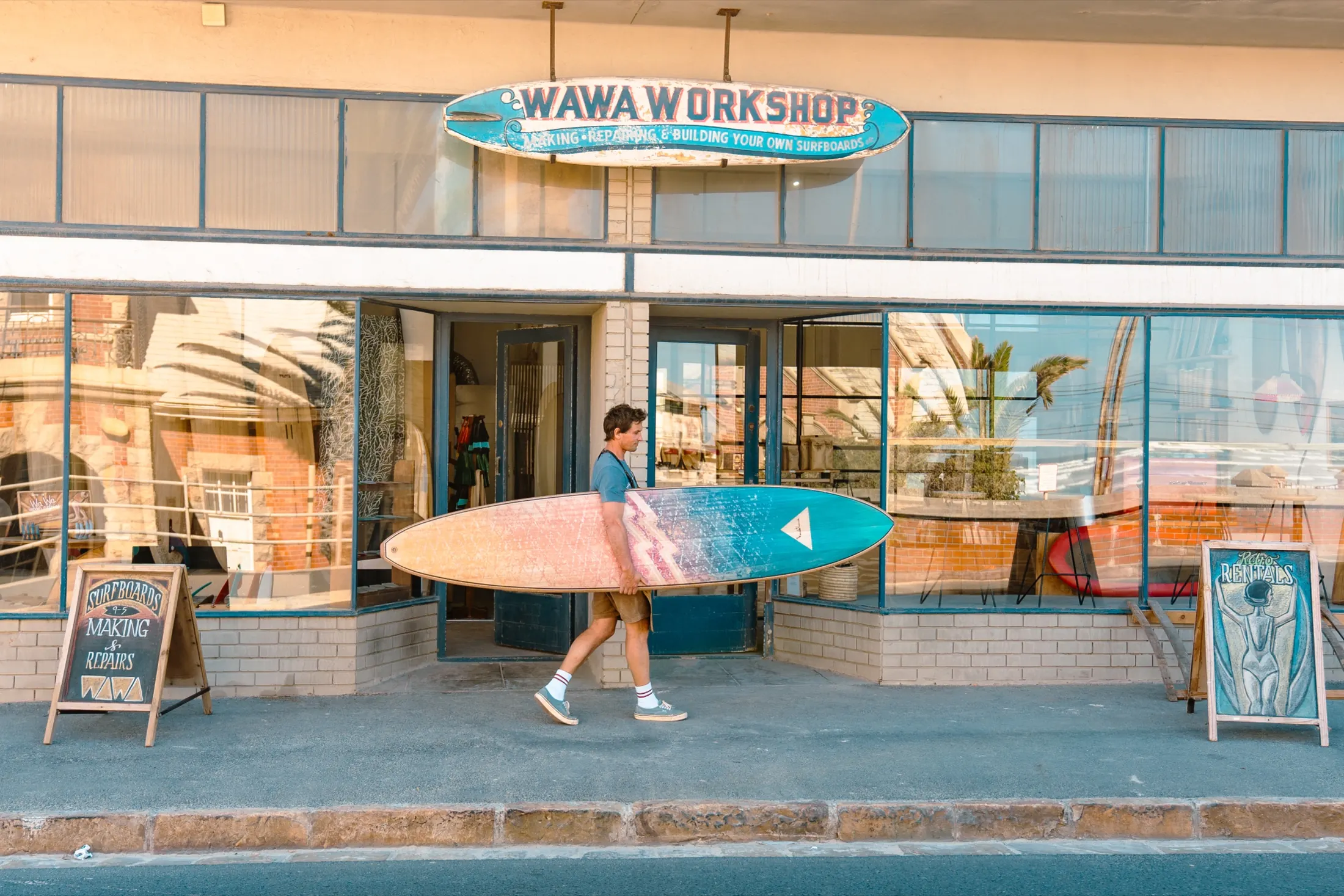 The Wawa Workshop shopfront in Muizenberg, a surfer walking past with a wooden board
