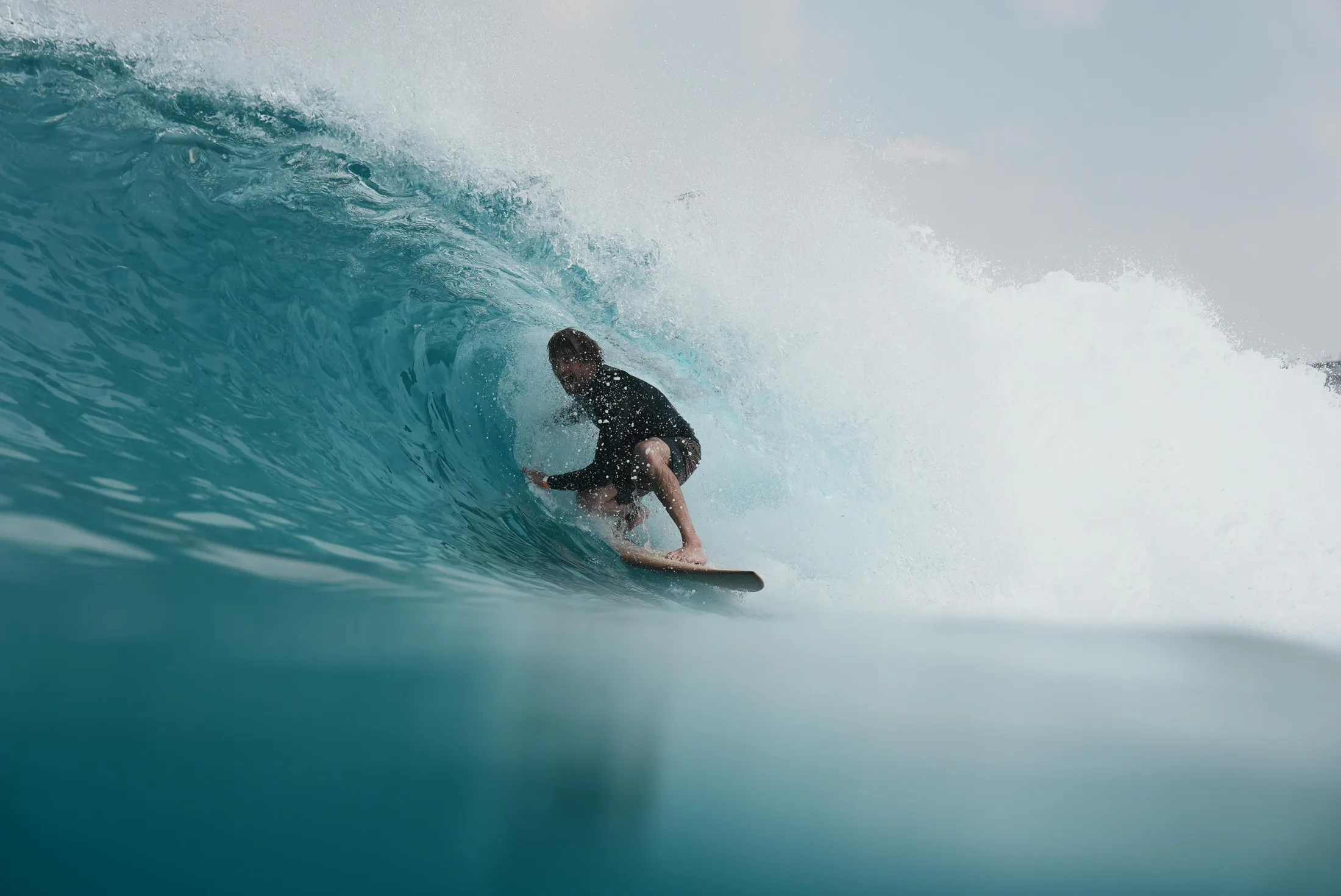 A surfer crouched inside a teal barrel on a wooden board, shot from the water