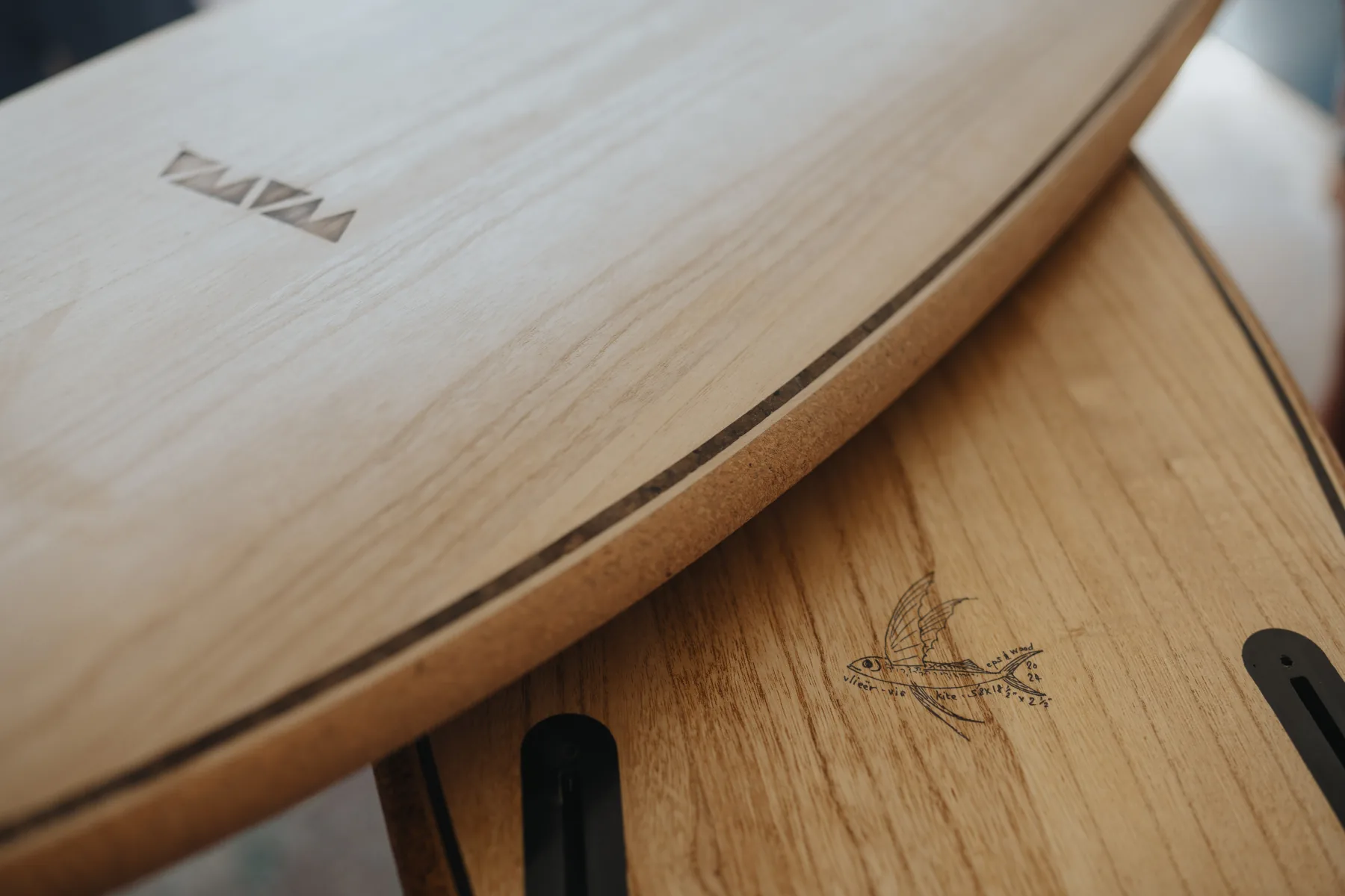 Two paulownia boards stacked, showing the WAWA deck inlay, a cork rail with a dark pin line, and a hand-drawn flying-fish stamp