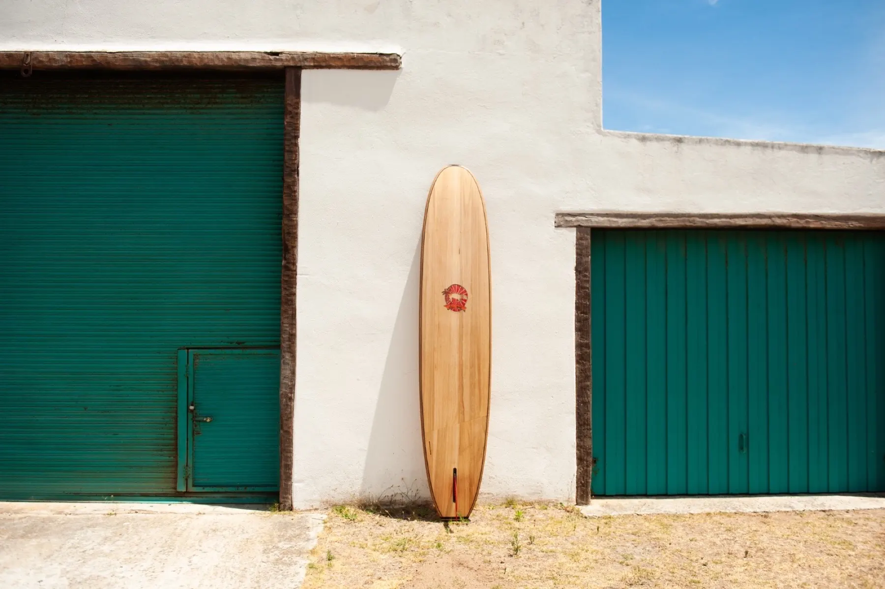 A paulownia longboard leaning against a whitewashed wall between green garage doors