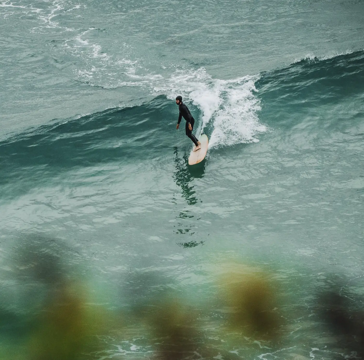 A surfer trimming across a green wall, seen from the cliff above