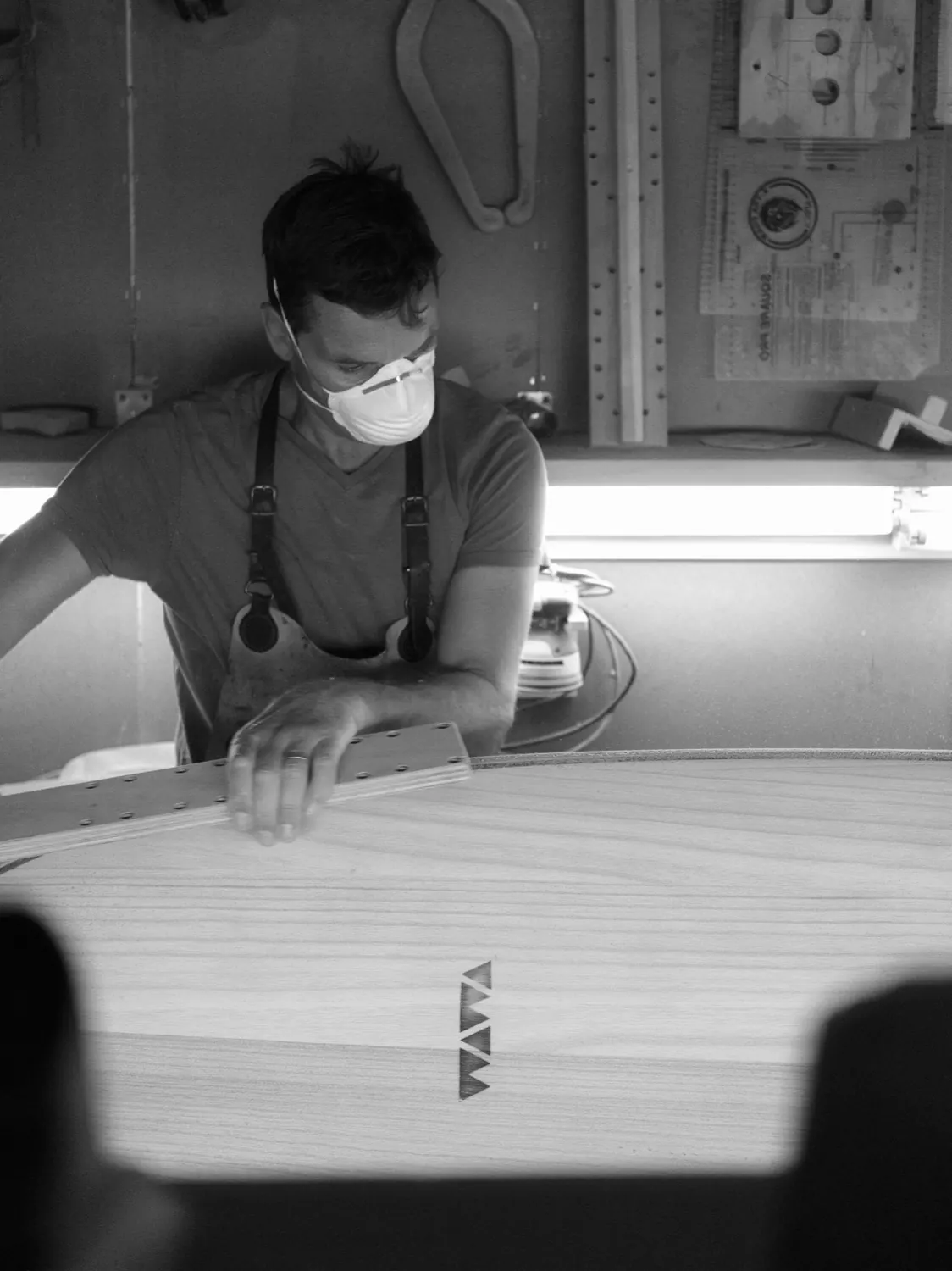 A shaper in a dust mask sanding a paulownia deck under a workshop window