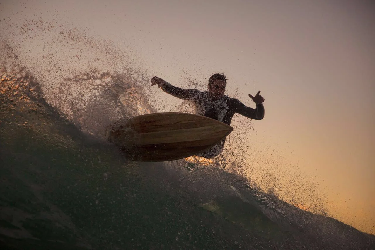 A surfer coming off the lip on a timber board at sunset, backlit through the spray
